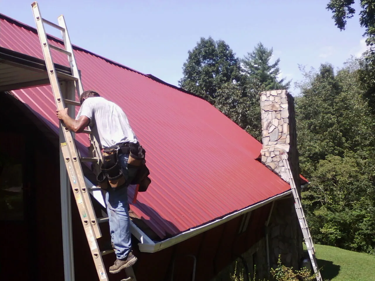 Roofer on a ladder beside a red metal roof and stone chimney — Soffit & Fascia Repair by Roofing Brothers in Park